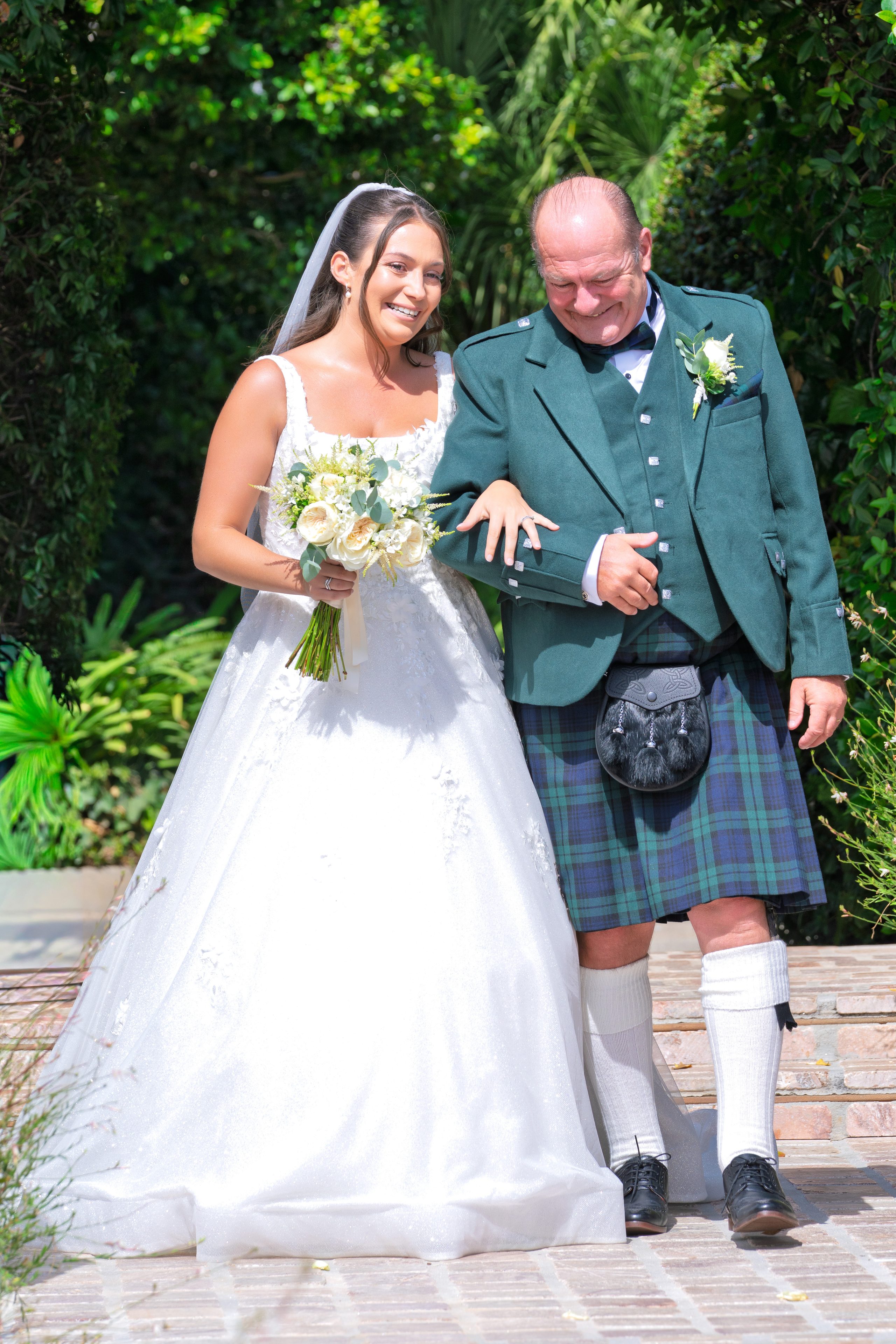 La novia, riendo, del brazo de su padre, con kilt, camino de la ceremonia — Cortijo Bravo, Vélez-Málaga