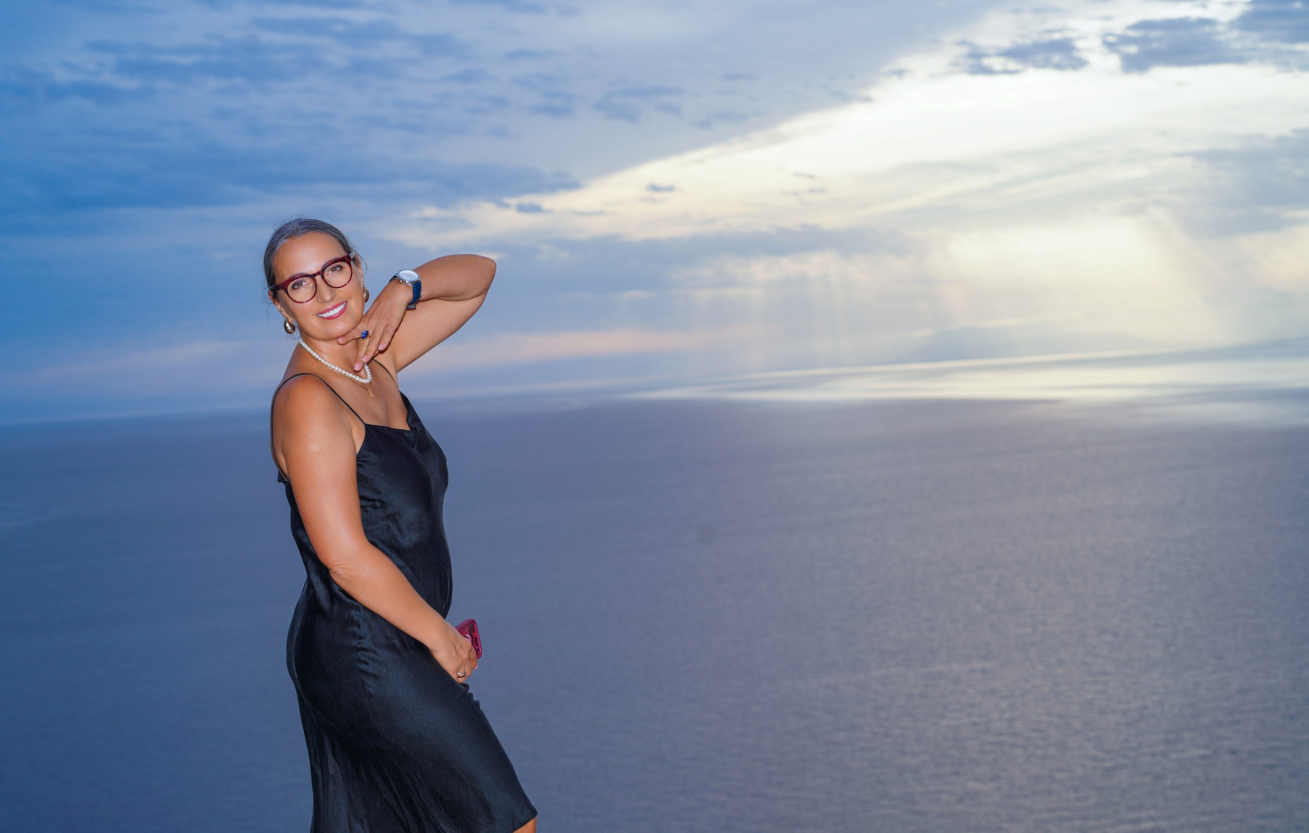 Woman in a black slip dress posing on a clifftop above the sea, sun rays breaking through the clouds — Cerro Gordo, La Herradura