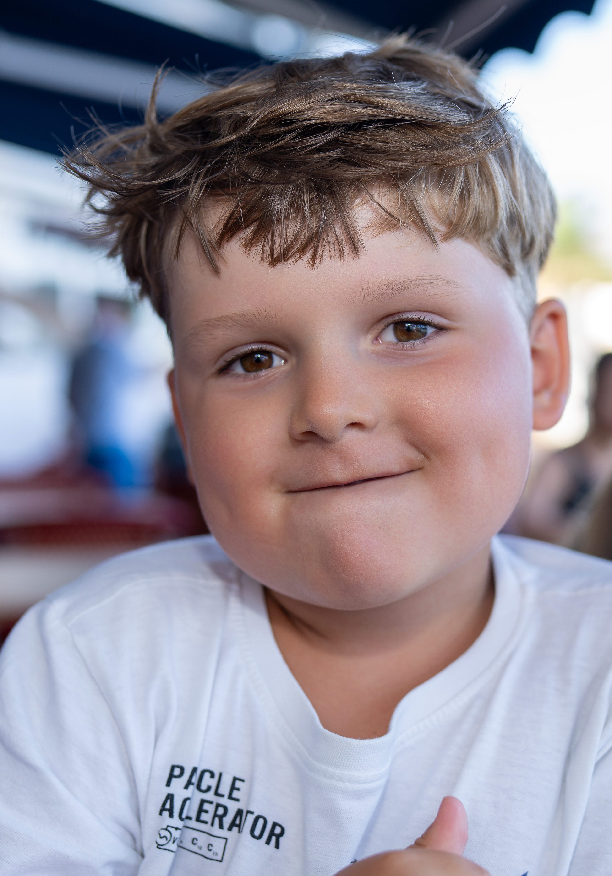 Close-up of a smiling boy with tousled fair hair in a white t-shirt, a blue awning blurred behind him