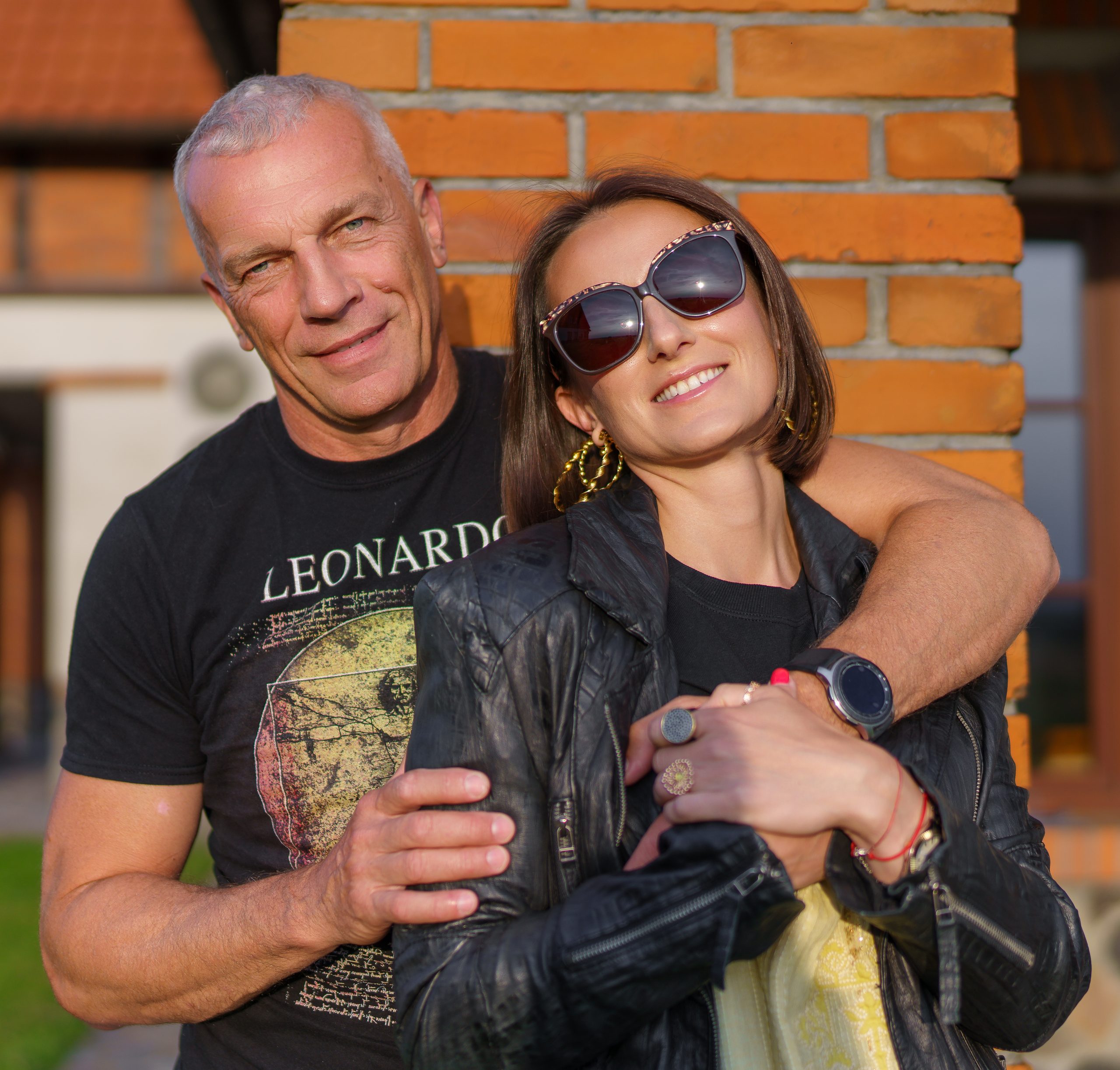 Homme en t-shirt imprimé enlaçant une femme à lunettes de soleil près d'un mur de briques